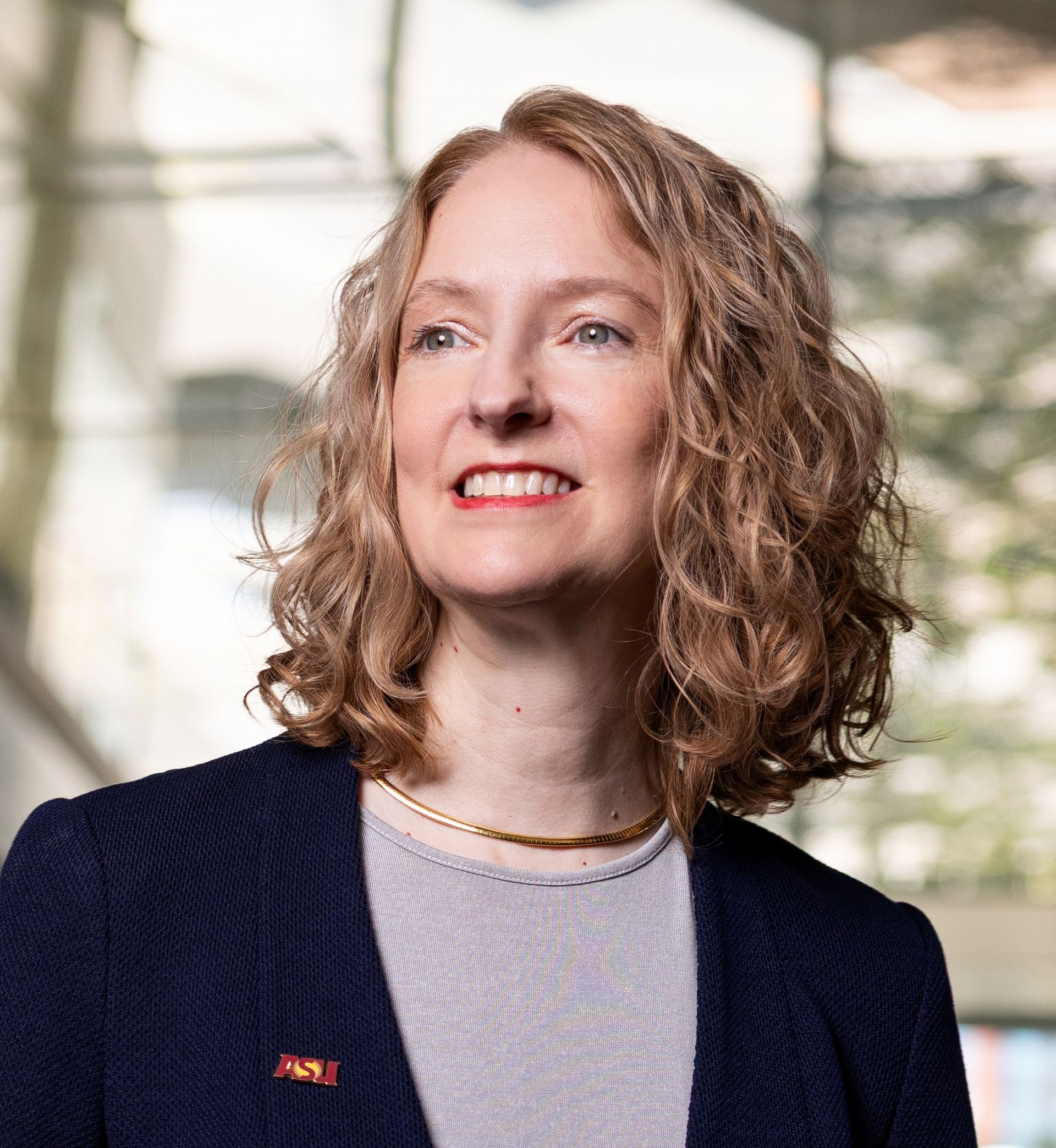 Portrait of a smiling woman with curly hair in a navy blazer indoors, lit by soft window light