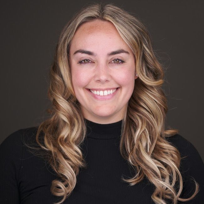 Smiling woman with wavy blonde hair wearing a black top on a dark background