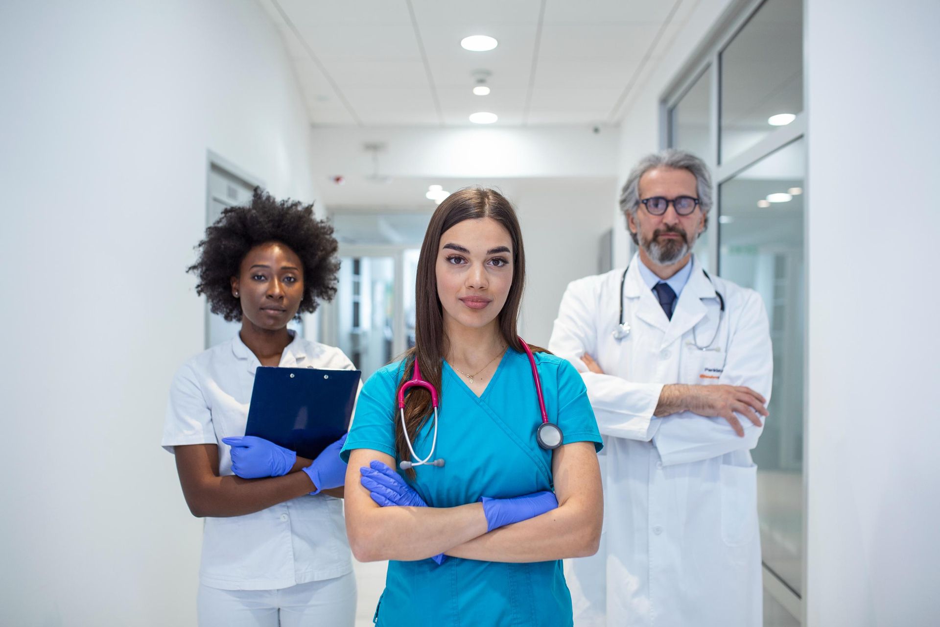 Three medical professionals standing in a bright hospital hallway, one holding a clipboard and one in scrubs with a stethoscope.