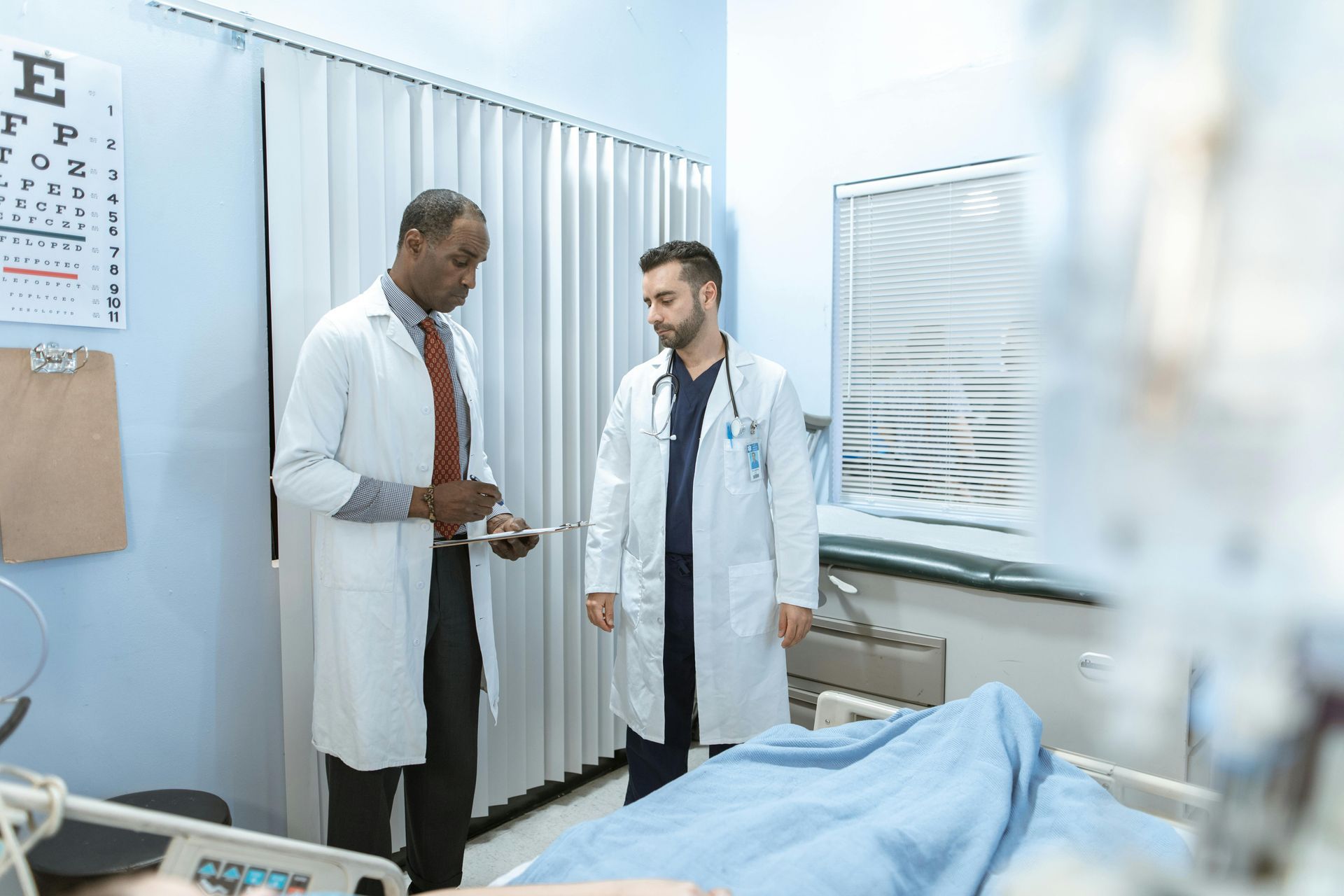 Two doctors in white coats confer in a hospital room beside a covered patient bed.