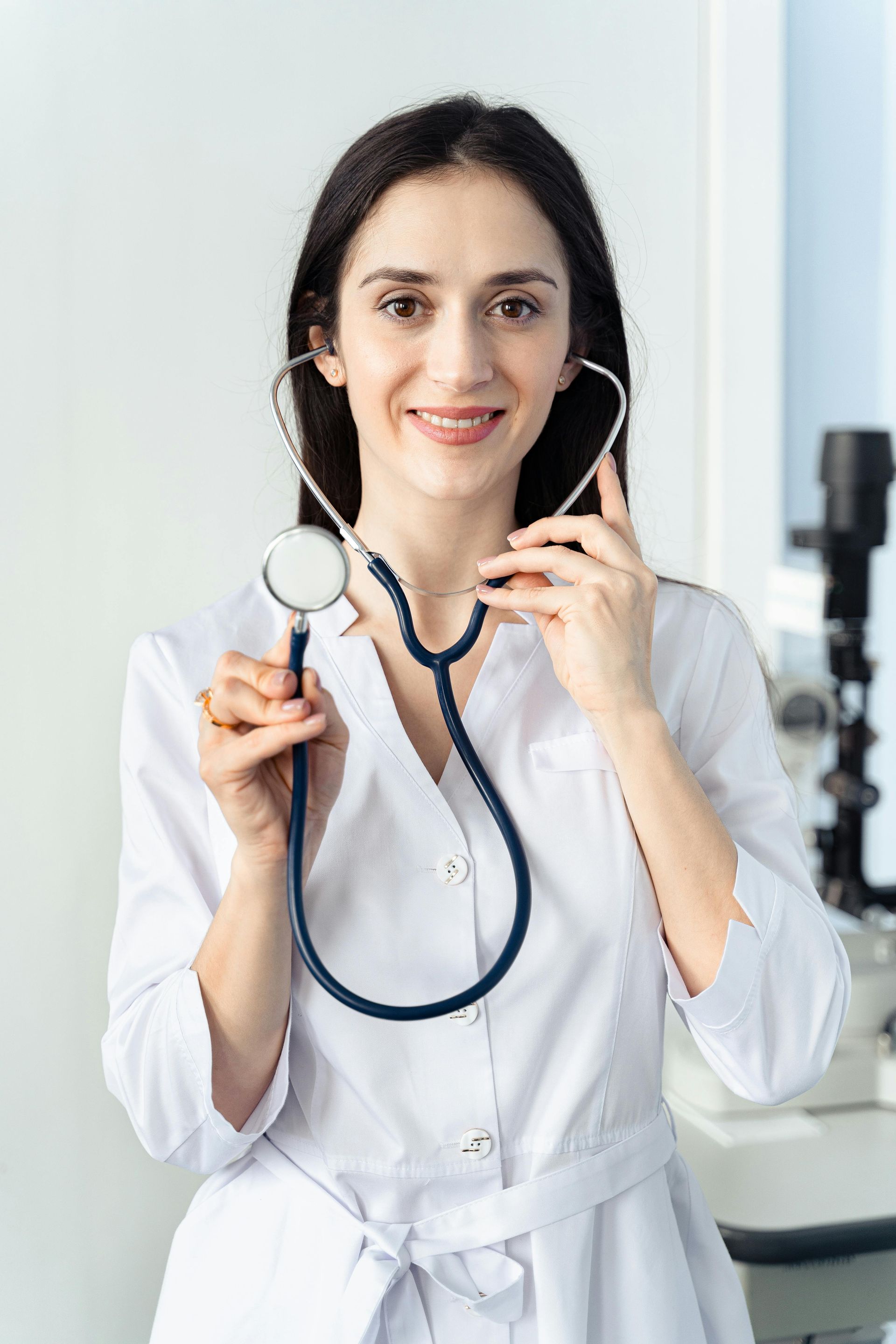 Doctor holding stethoscope in a clinic, smiling at the camera, wearing a white coat.