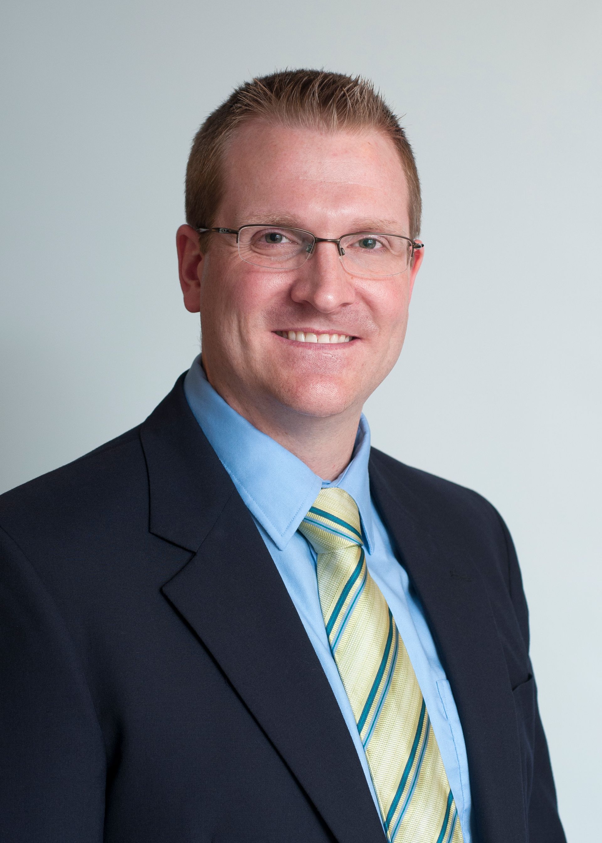 Professional headshot of a smiling person in a dark suit, blue shirt, and striped tie against a light background