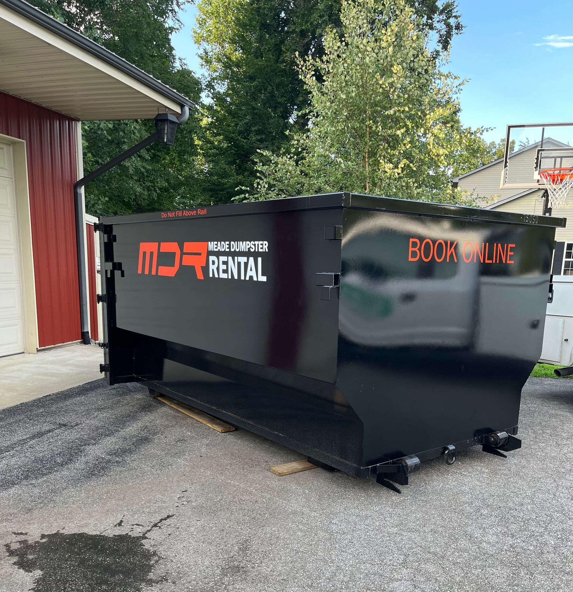 Black rental dumpster beside a building on gravel, with orange lettering and a basketball hoop in the background