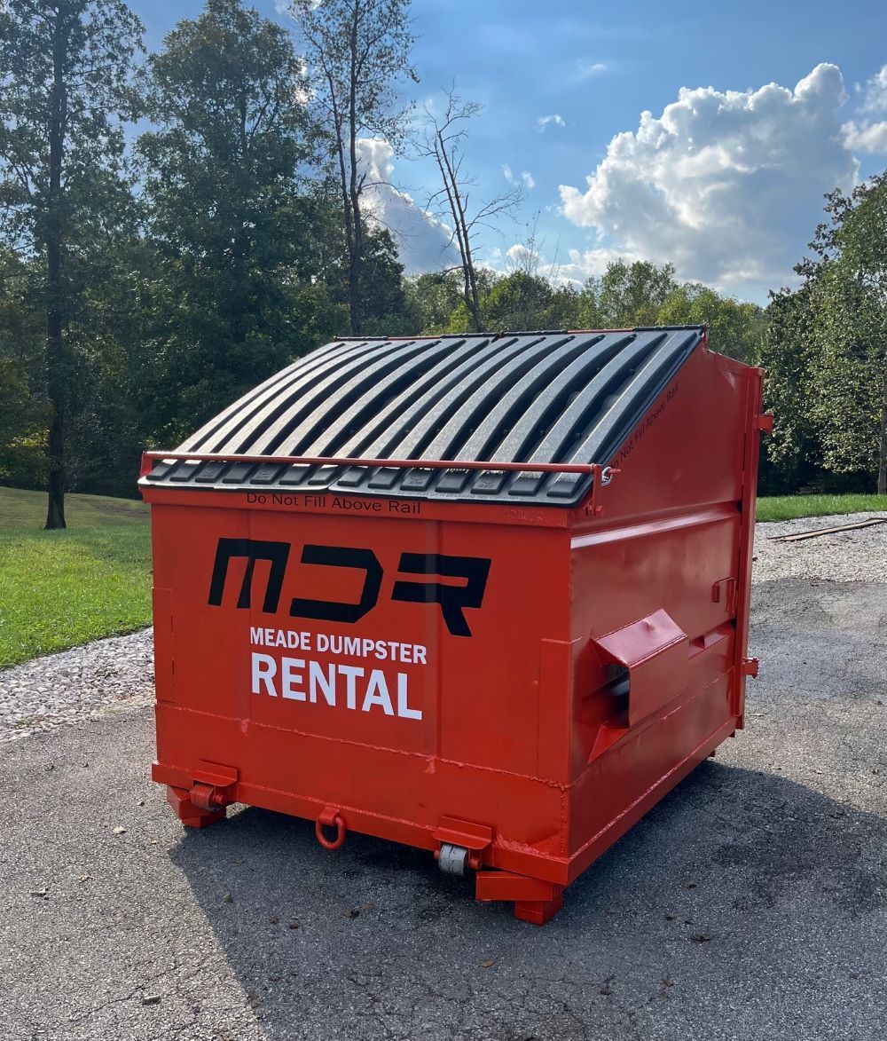 Red rental dumpster with black lid on gravel beside trees and blue sky