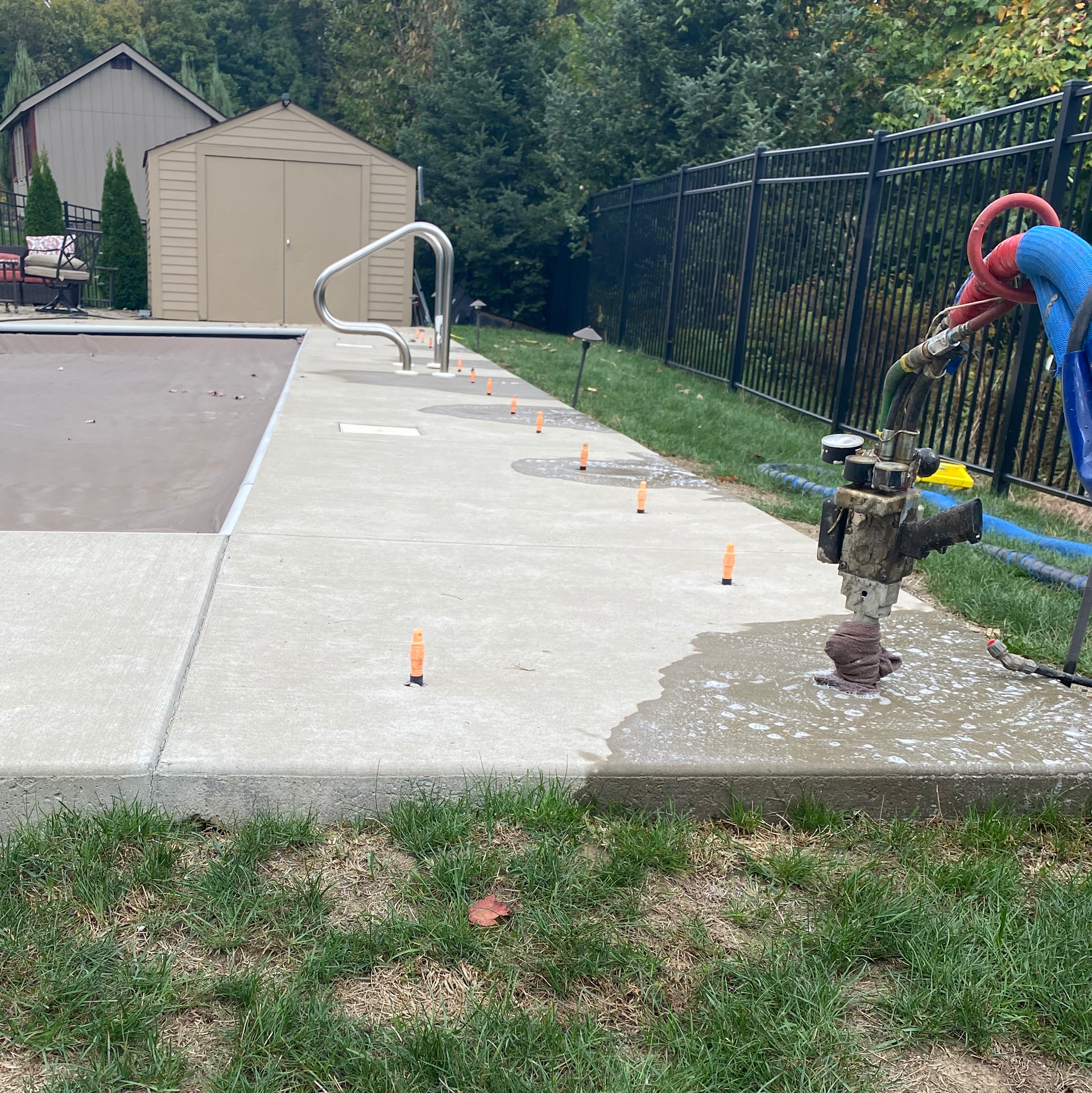 Poolside concrete repair: person uses equipment near a water valve. Orange markers, shed, black fence, and grass surround.