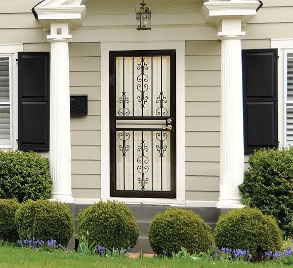 White front door with black iron screen, flanked by columns, shutters, and trimmed shrubs at a house entrance