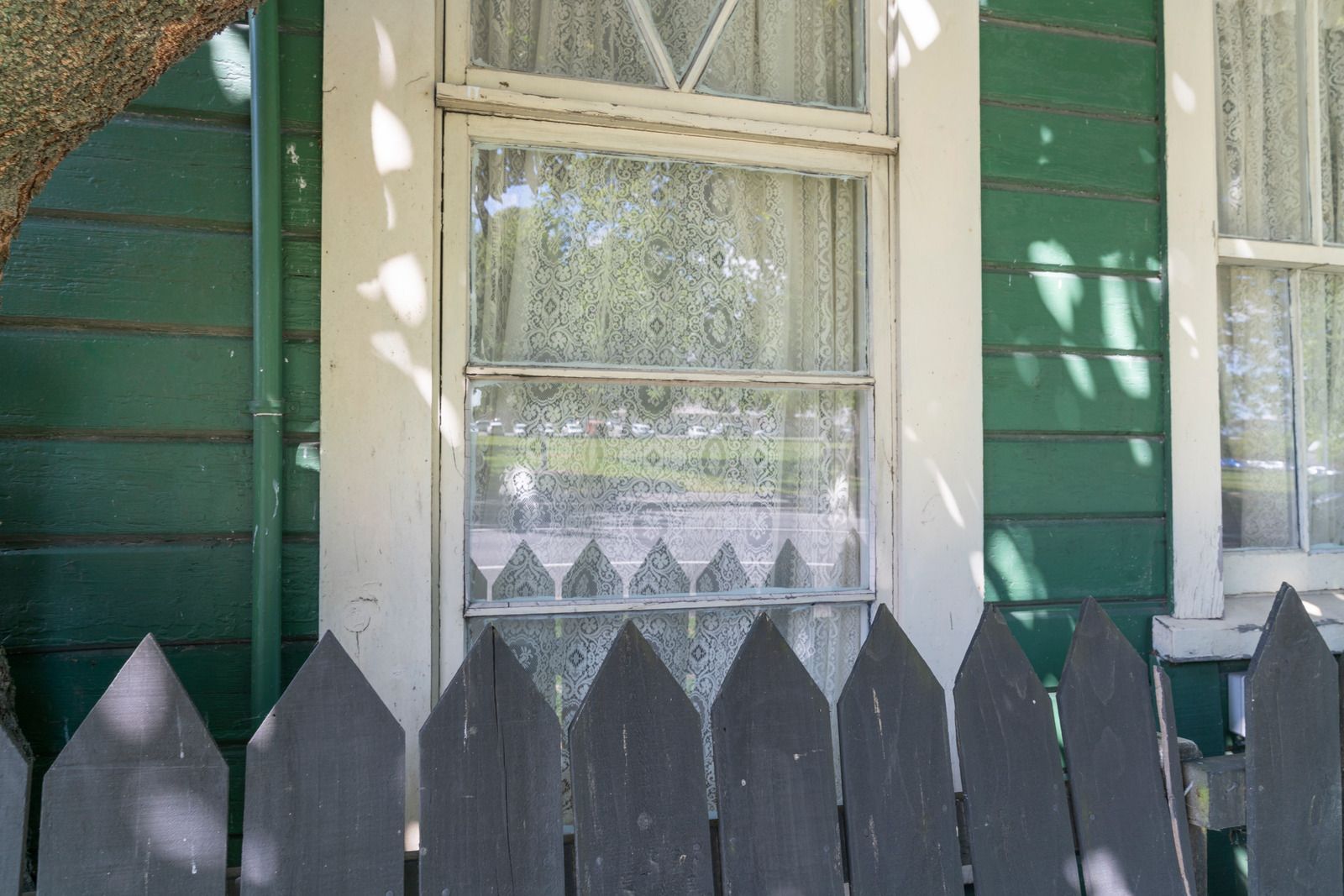 Green house window with white trim and lace curtain behind a dark picket fence