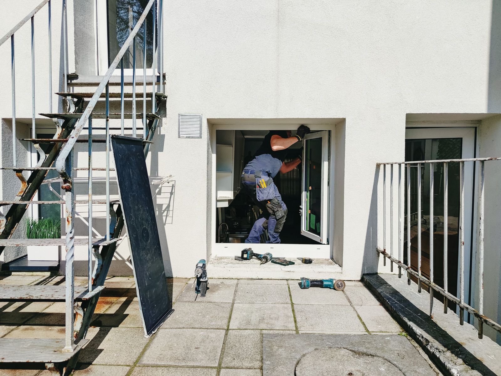 Worker entering a small window opening at a building exterior, with tools and a ladder nearby.