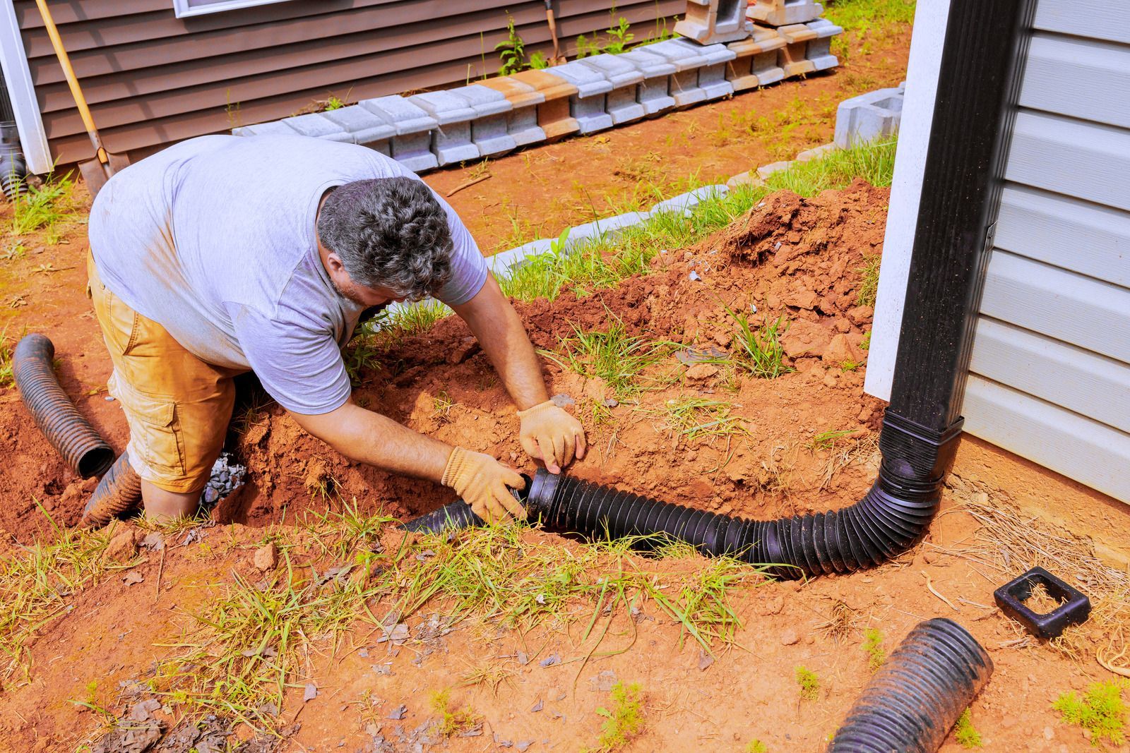 Worker installing black drainage pipe in a muddy yard beside a house