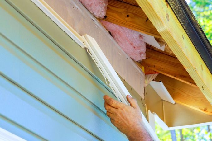 Hand installing white vinyl siding beneath a roof eave on a house exterior