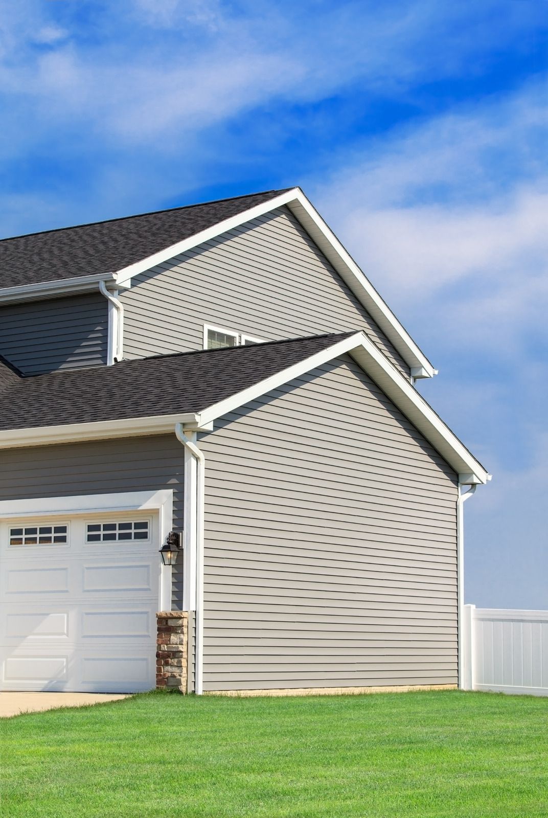 Suburban house with white siding, dark roof, garage, and green lawn under a blue sky