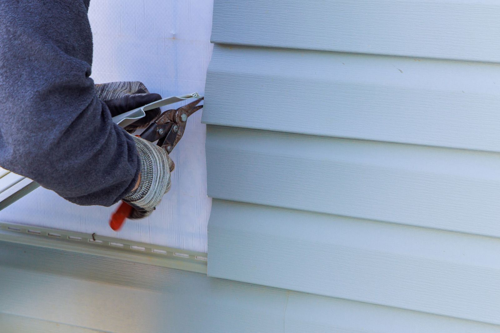 Gloved worker using a scraper to remove white siding from a house exterior