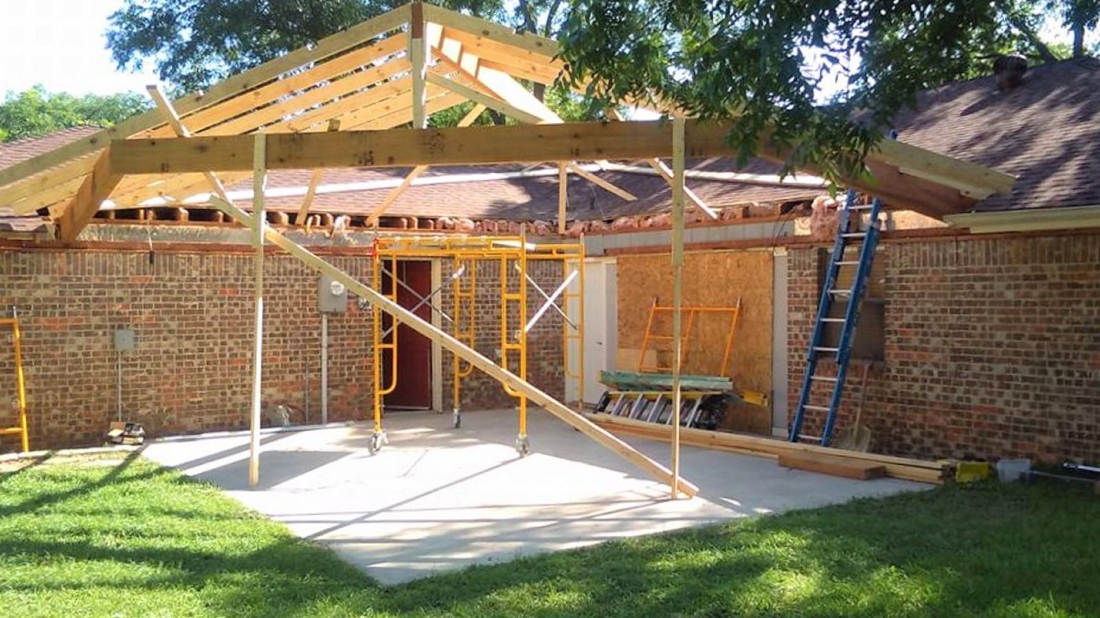 Partially built house addition with exposed wooden framing, brick walls, and ladders in a sunny yard