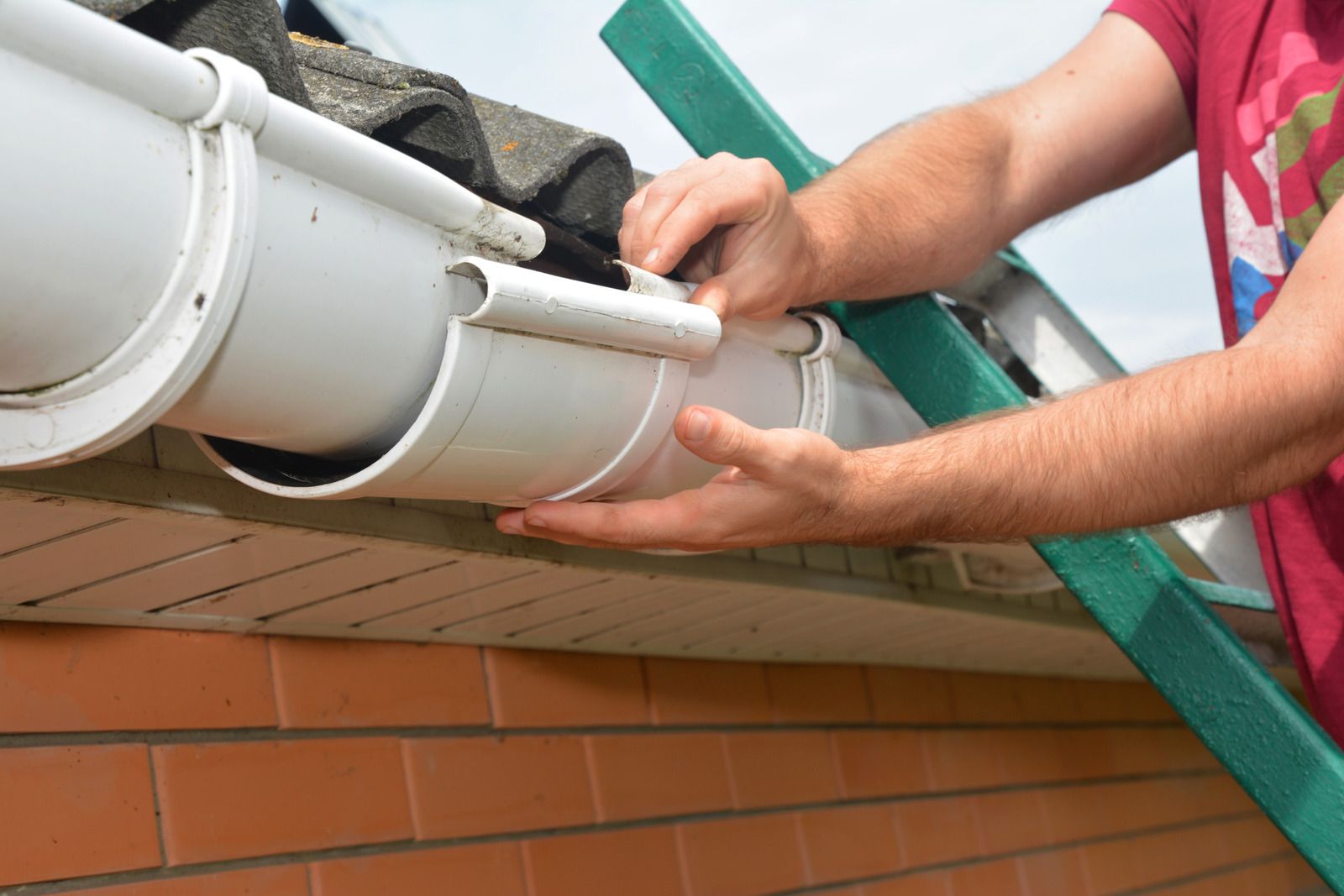 Hands installing a white gutter on a brick house roofline with a green ladder nearby