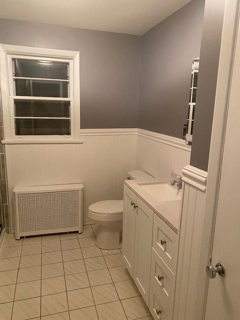 Bathroom with white wainscoting and cabinets, gray walls, a window, and a white tiled floor.