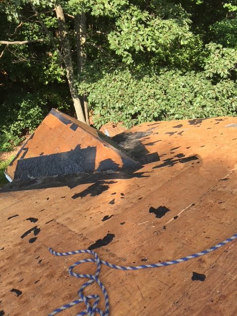 Rooftop with damaged shingles; dark patches near a chimney, and a blue rope in the foreground. Trees in the background.