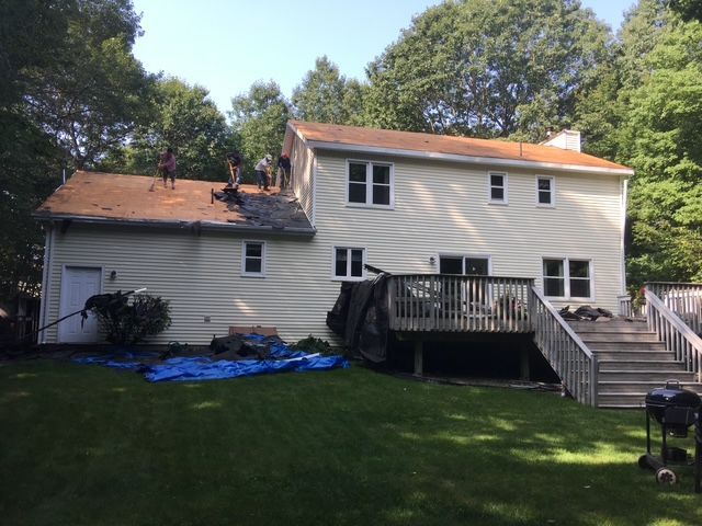 Roofers working on a residential house with a deck. Blue tarp on the lawn; trees in the background.