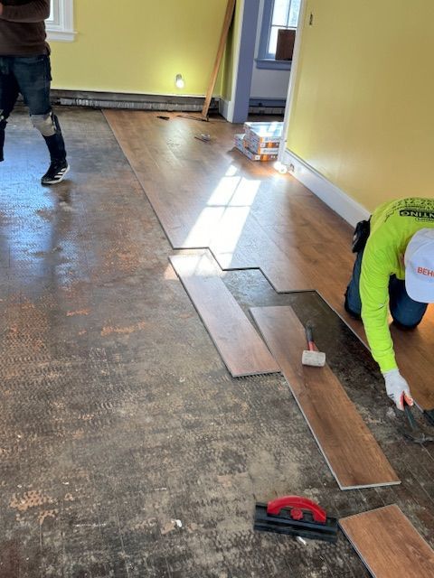 Two workers installing hardwood flooring in a room with yellow walls. One kneels, placing planks.