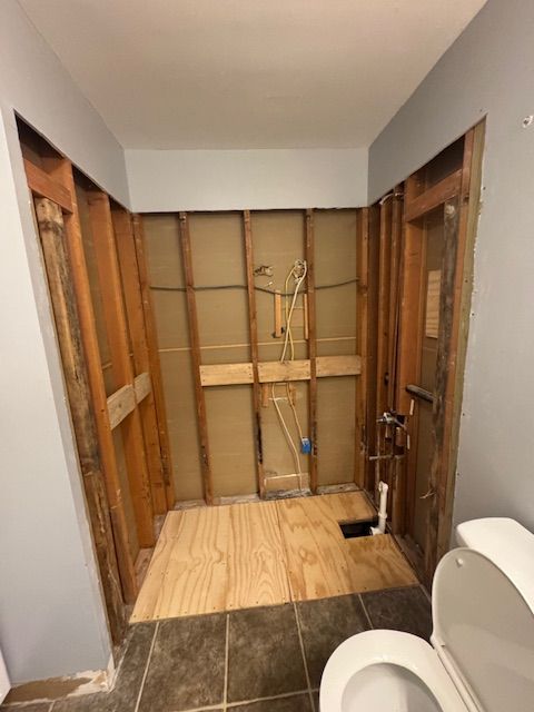 Bathroom under renovation, studs exposed. Plywood floor, plumbing visible on the center wall. Toilet in the foreground.