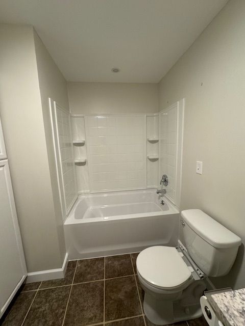 Bathroom with white tub, toilet, and shower surround. Brown tile floor. Light walls.