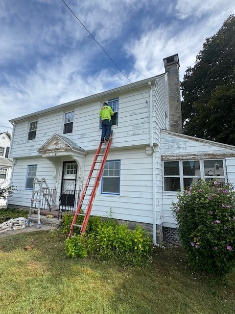 Person on ladder painting the side of a white house. Cloudy sky, green grass, and bushes visible.