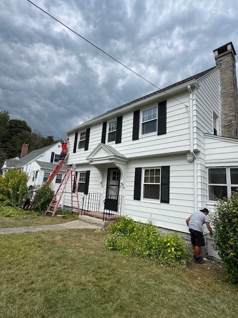 Two-story white house with black shutters, people working on exterior under cloudy sky.