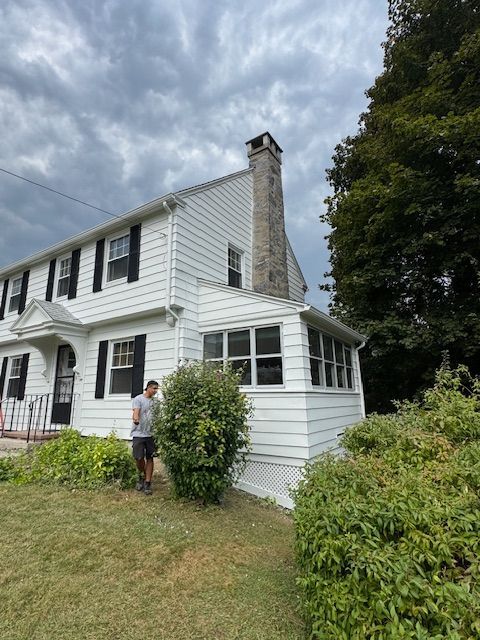 White house with black shutters, chimney, person near bushes on a cloudy day.