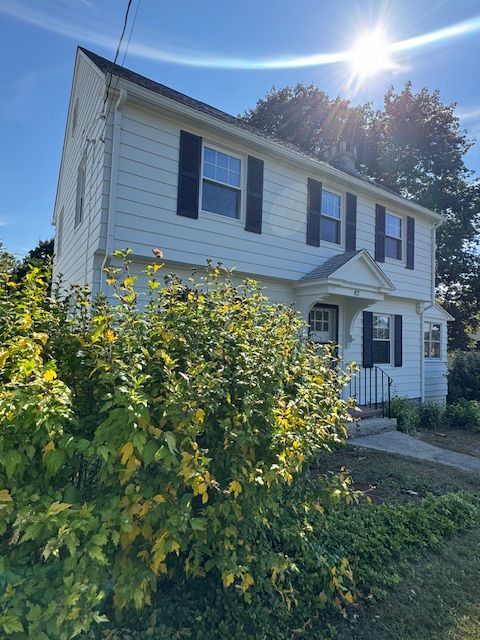 White two-story house with black shutters, door, and a sunlit sky. Bushes and greenery in front.