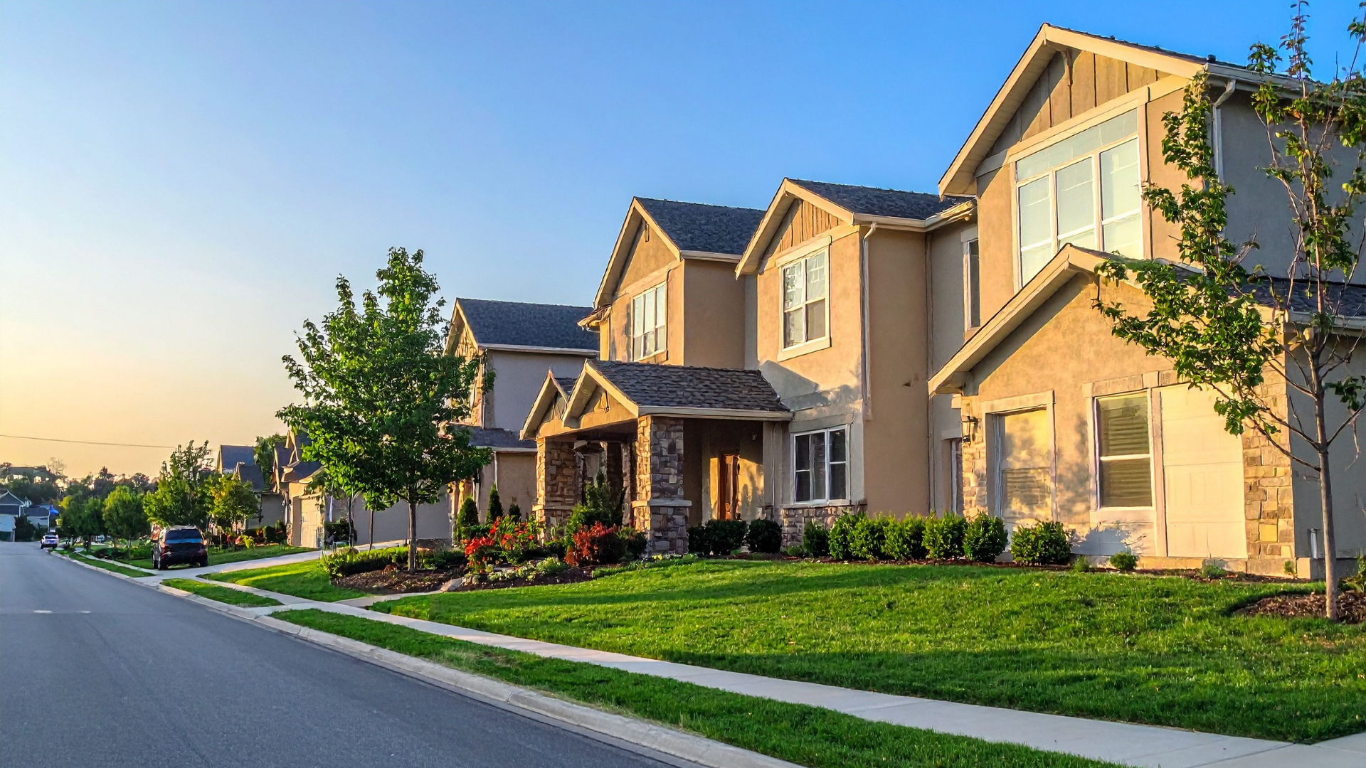 Row of beige houses with green lawns and trees on a sunny street.