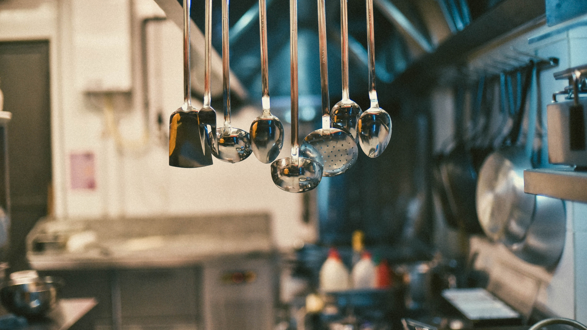 Kitchen utensils hanging in a commercial kitchen, blurred background with stainless steel surfaces.