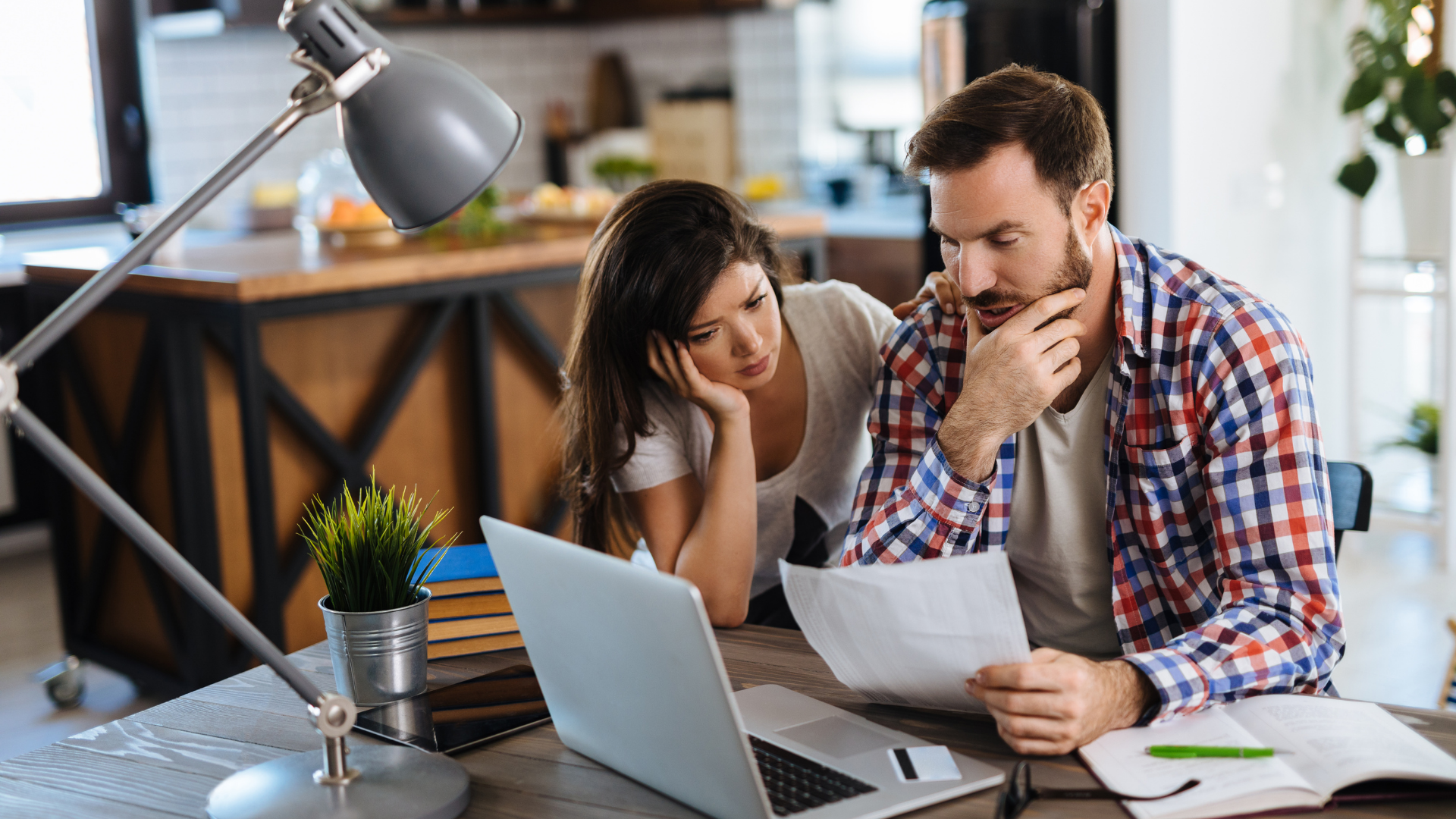 Couple reviewing paperwork at a desk, looking concerned, laptop open, lamp lit.