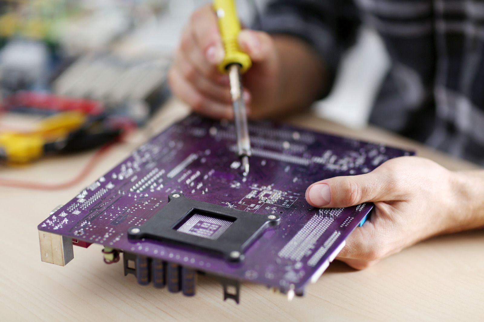 Hands using a screwdriver to repair a purple circuit board on a workbench