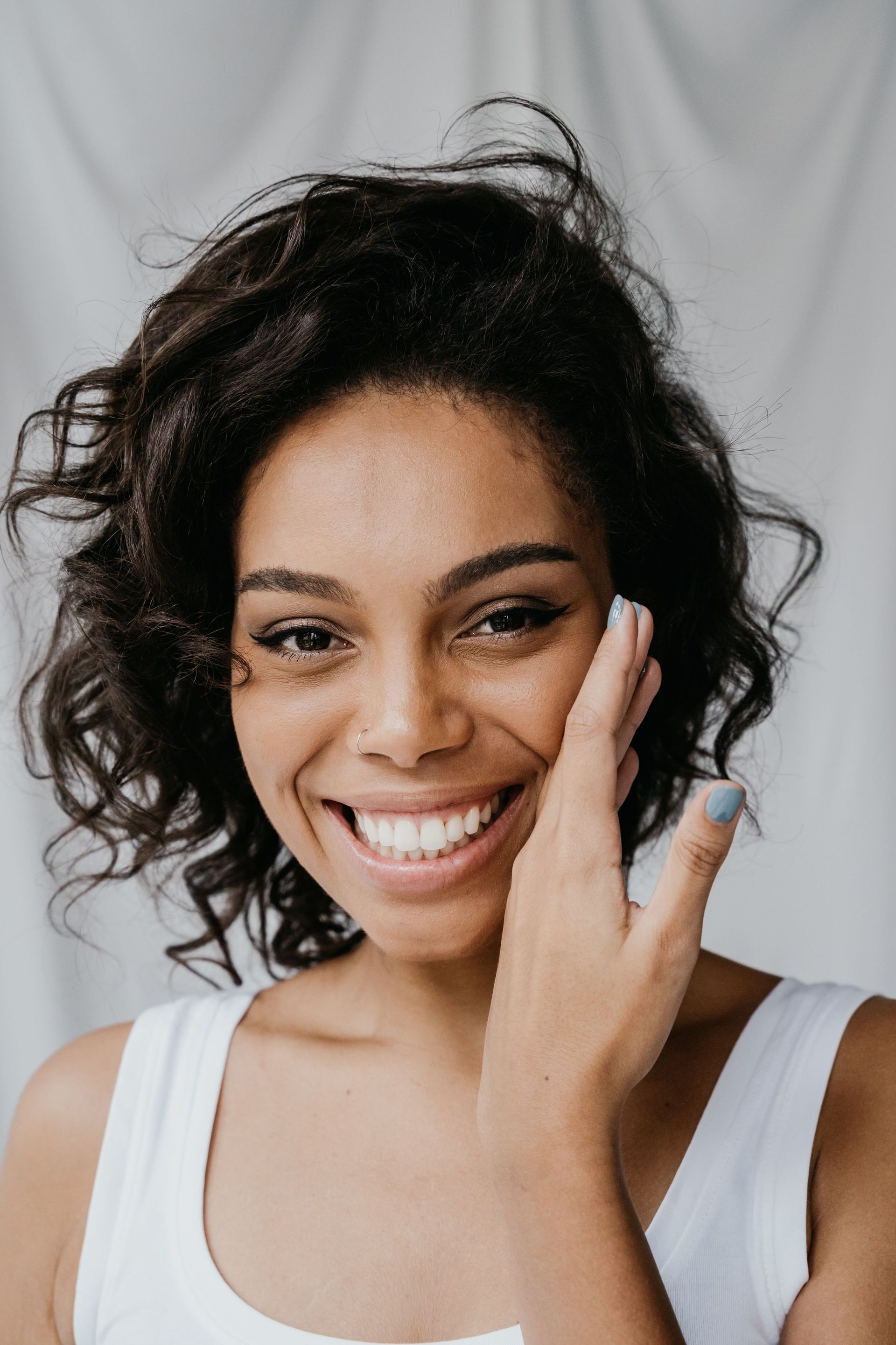 Smiling person with curly hair touching their cheek against a light gray background