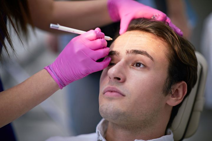 Person having eyebrows filled in with a pencil while wearing a white paper face shield