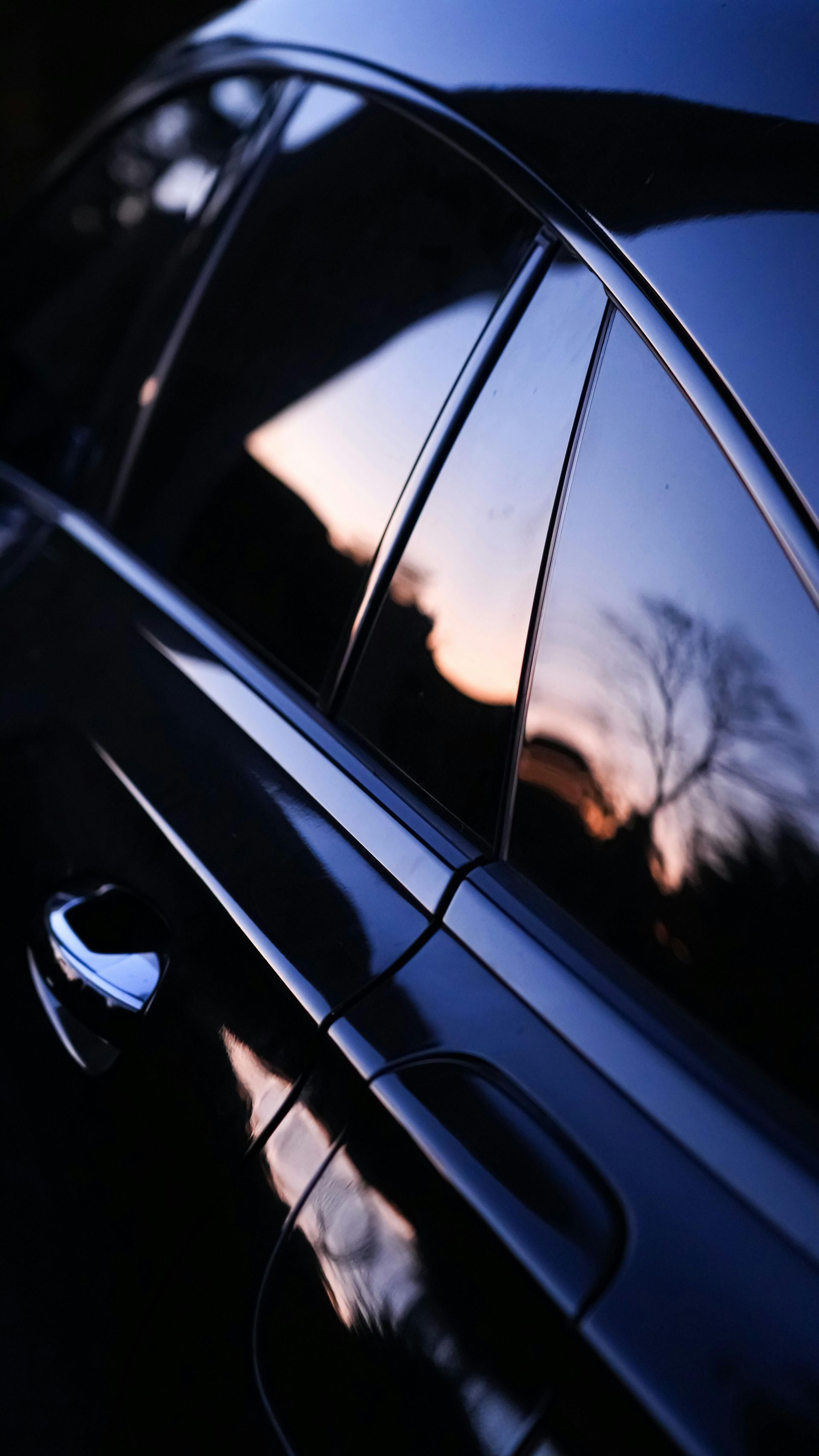 Close-up of a glossy black car door reflecting a sunset sky and silhouetted trees