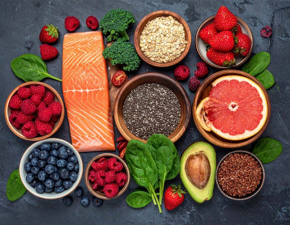 Assortment of fresh, healthy foods in bowls: salmon, berries, spinach, broccoli, avocado, quinoa, chia seeds, and grapefruit.
