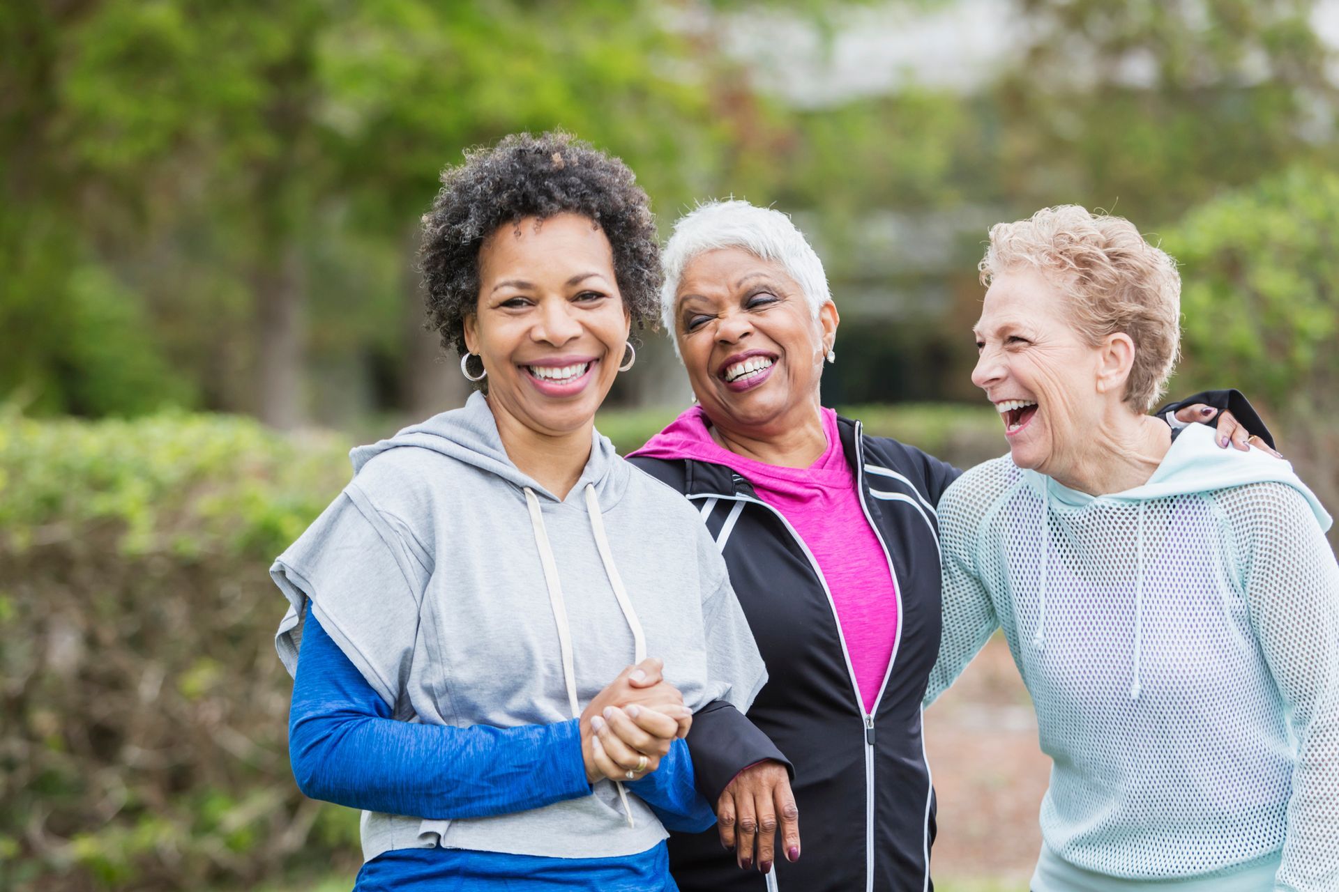 Three older women are standing next to each other in a park and laughing.