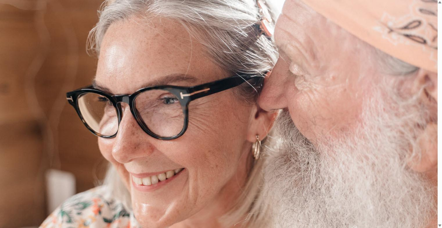 Older bearded man leaning close to whisper to a smiling older woman wearing glasses