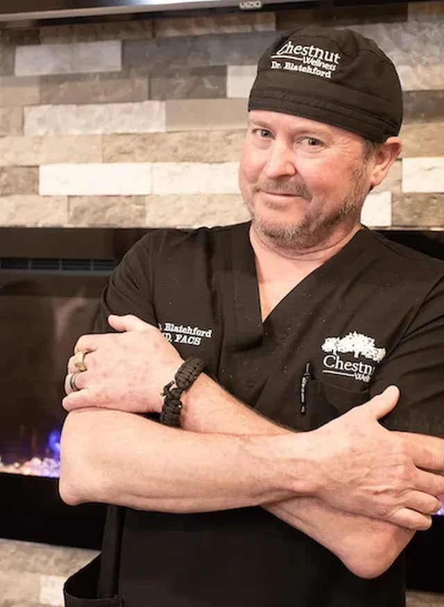 Doctor Blatchford in a scrub suit and hat is standing with his arms crossed in front of a fireplace .