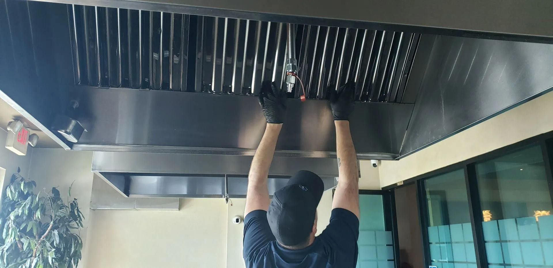 Person wearing gloves adjusts a stainless steel kitchen exhaust hood in a commercial setting.