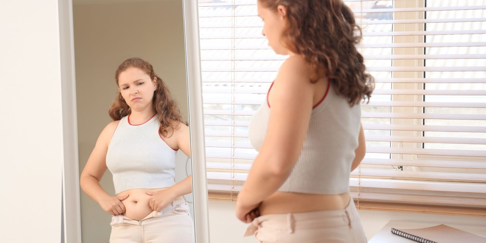 Person standing by mirror adjusting shirt in a bright room with window blinds