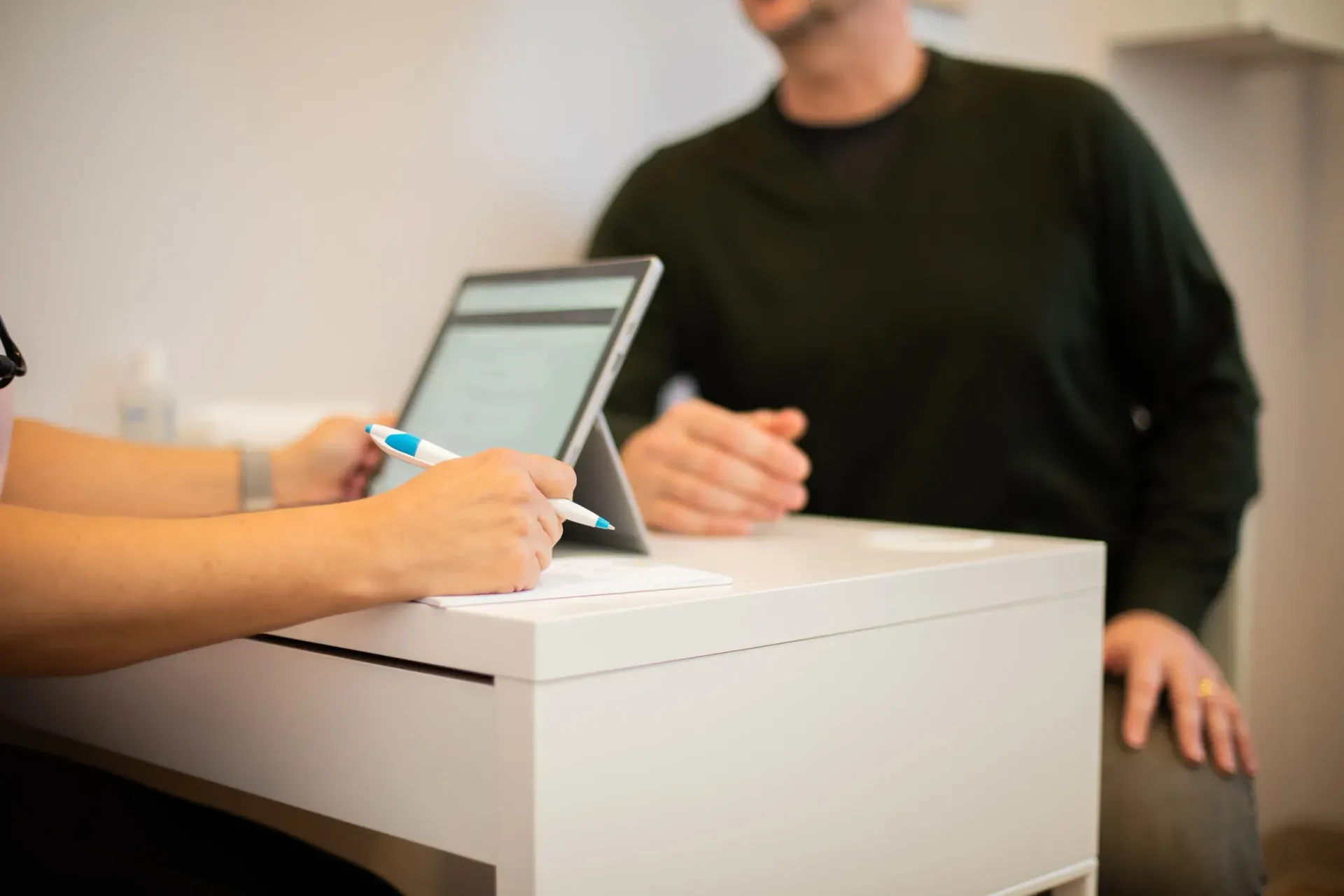Person using a tablet at a white counter while another person sits nearby in a blurred indoor setting