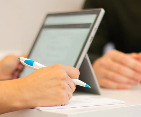 Hands holding a blue pen over a notebook beside a tablet on a desk in a meeting setting