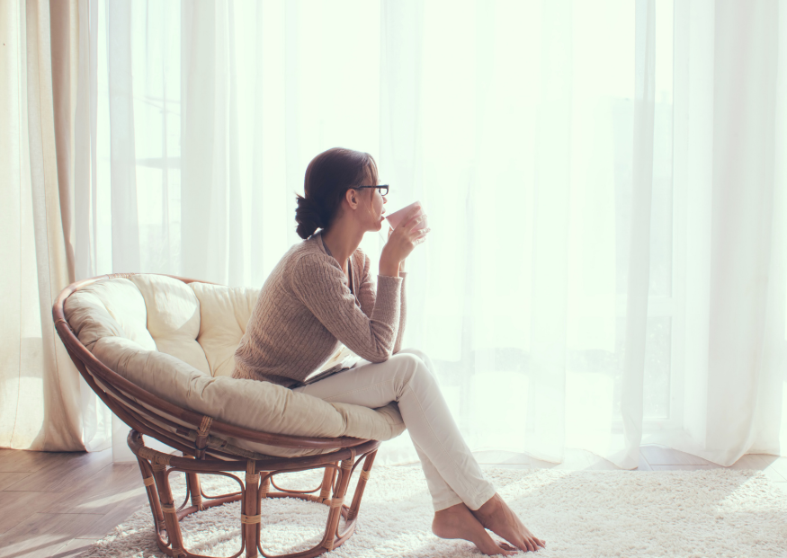 A person sits in a round rattan chair, wearing a sweater and pants, drinking from a mug while looking out a window.