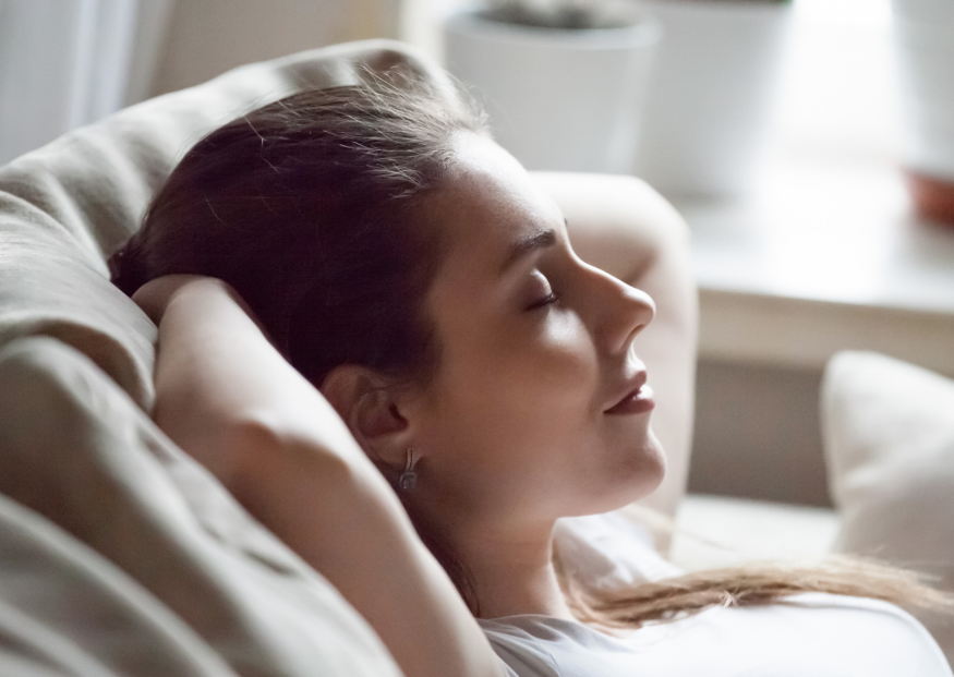 A person with their eyes closed, resting with their hands behind their head, leaning back on a soft couch.