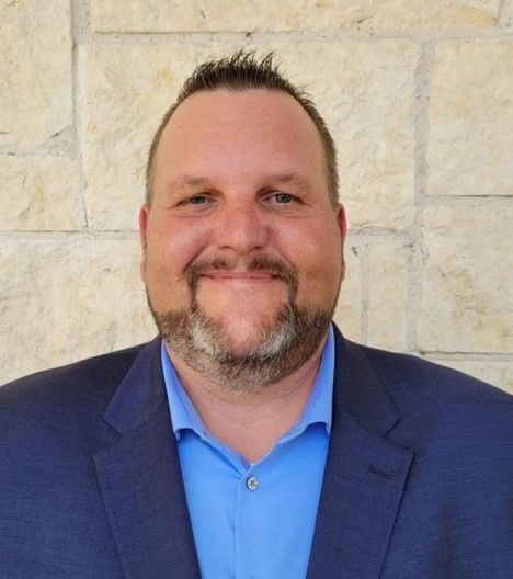 A man in a suit and blue shirt is smiling in front of a brick wall.