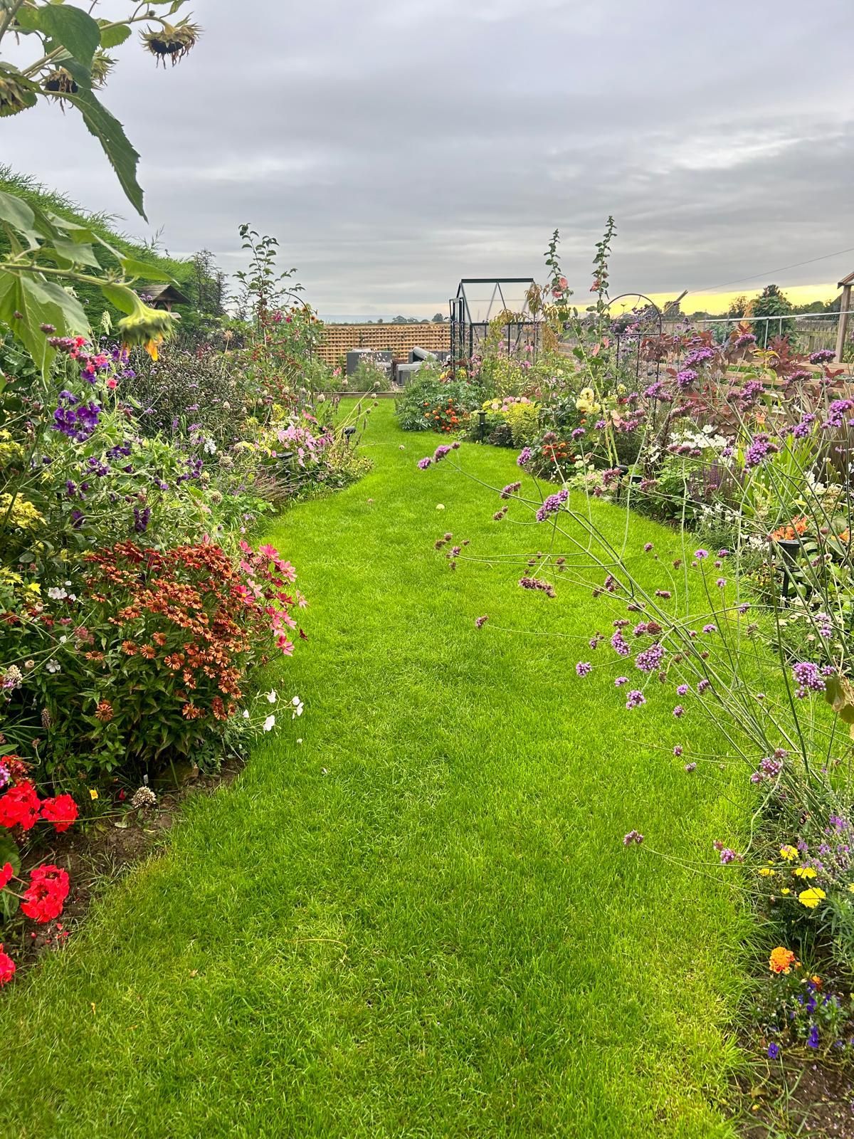 Lush green grass path through a colorful garden with various flowers under a cloudy sky.