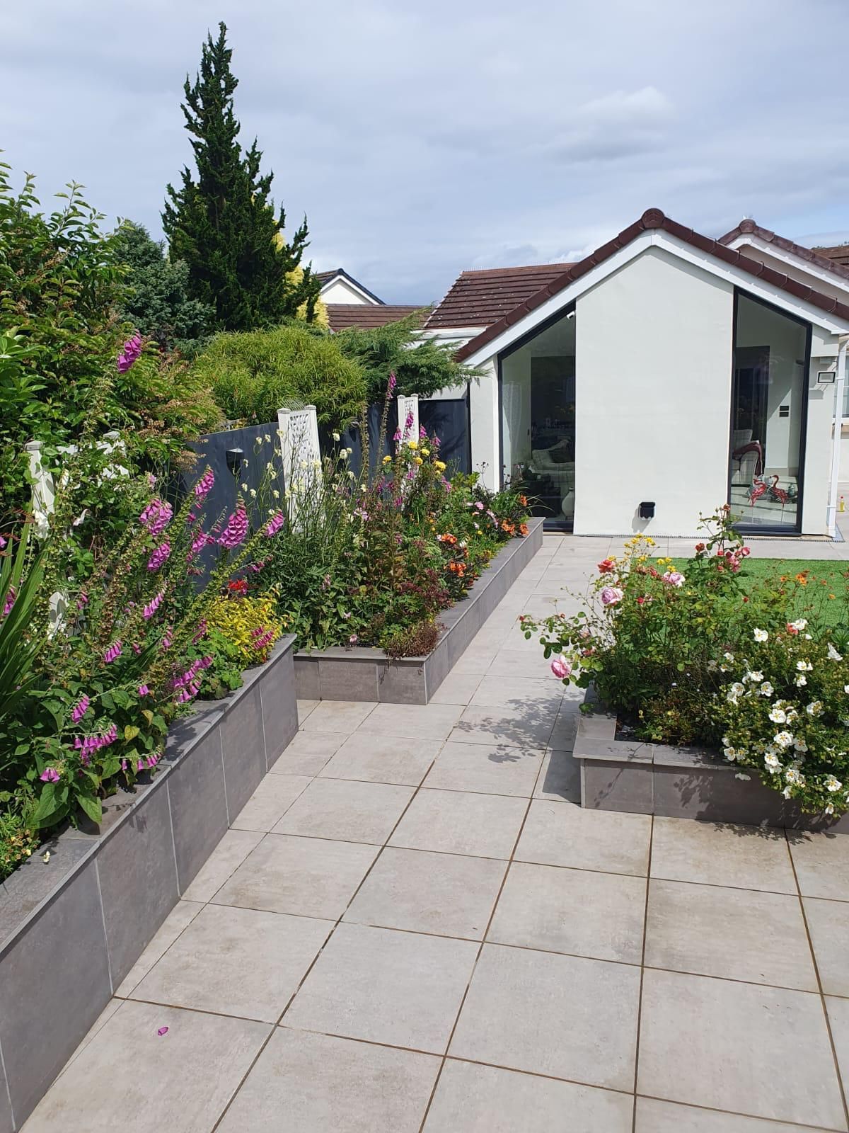 A sunny garden patio with flower beds and a white house in the background.