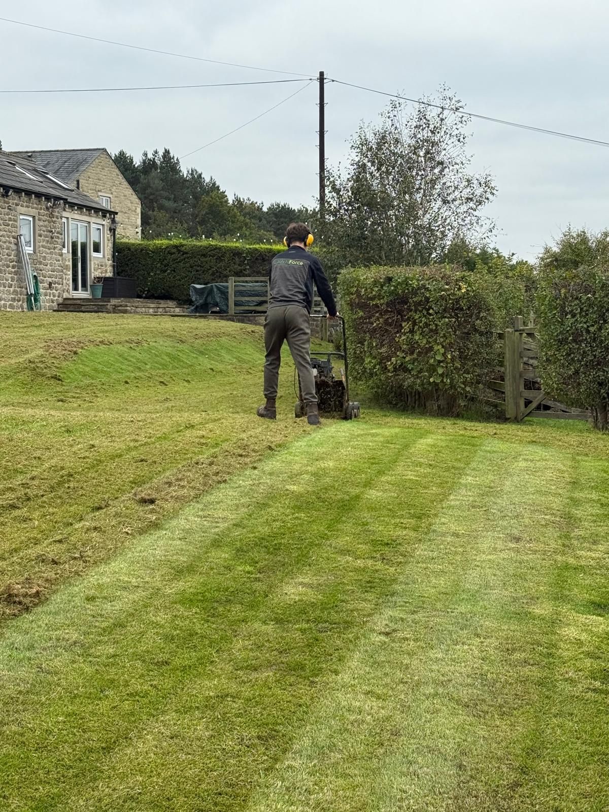 A person mowing a green lawn with a push mower; stone building and hedges in the background.