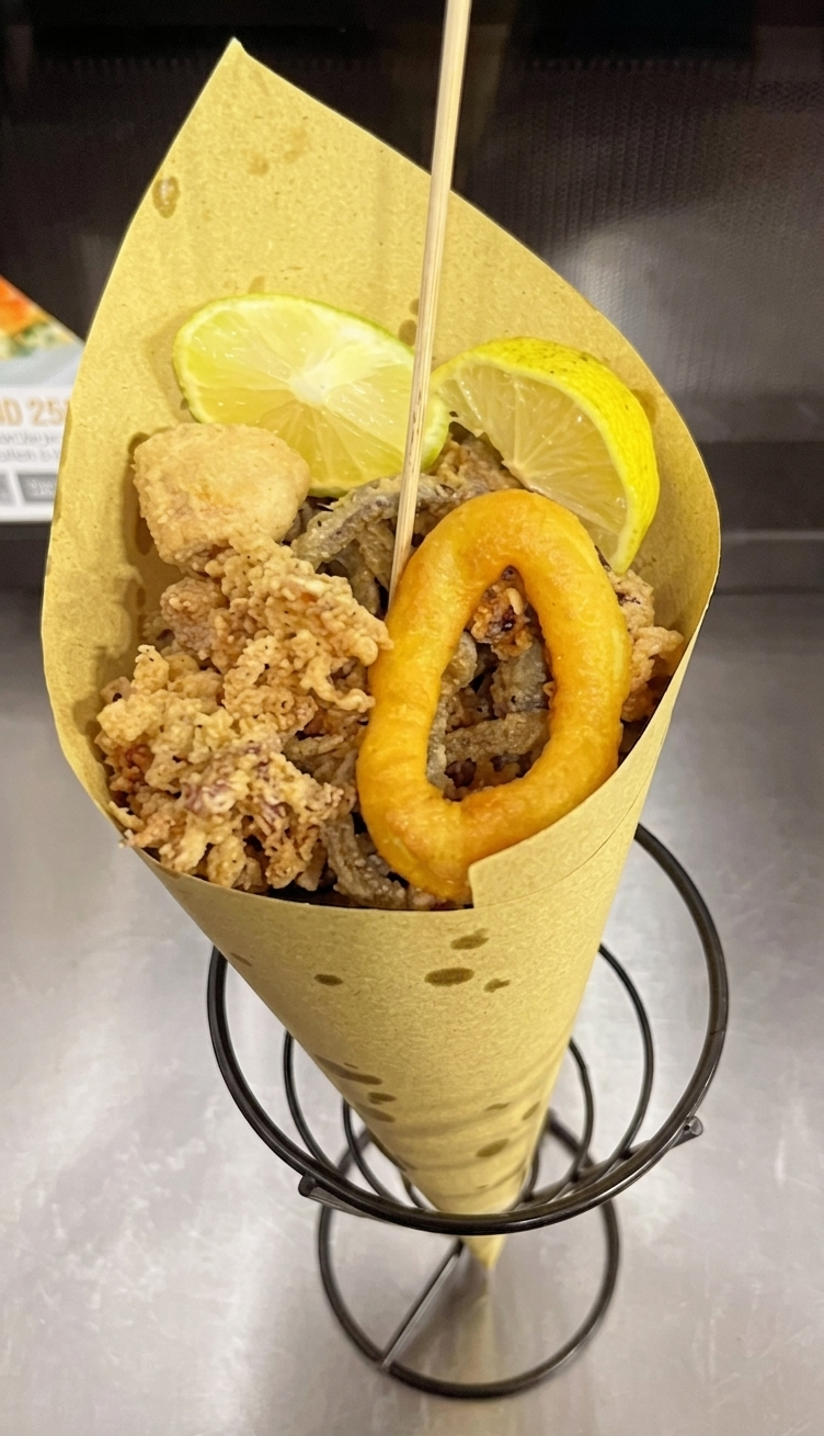 Cono di carta contenente snack fritti misti con spicchi di limone e anelli di cipolla, su un supporto di filo metallico.